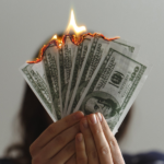 woman holding paper money in front of her face that is on fire - featured image for sunk cost effect blog by truby achievements