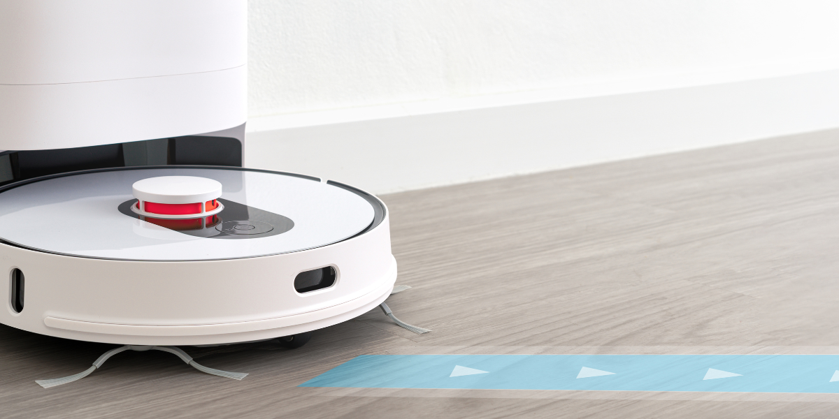 A white robotic vacuum cleaner docks itself at a charging station on a light wood floor, with a blue directional arrow path overlayed in front of it – featured image for Learning from Failure blog by Truby Achievements.
