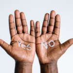 A hand hovers over two buttons—YES and NO—symbolizing the power of choice and conscious control. This image illustrates the concept of free won't and features the Achievement Nudge blog by Truby Achievements.